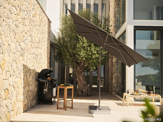ein_grossflaechiger_quadratischer_sonnenschirm_von_glatz_mit_stabilem_fuss_schuetzt_einen_modernen_grillplatz_der_sich_auf_der_terrasse_eines_modernen_hauses_befindet_begrenzt_von_einer_hohen_steinernen_mauer_auf_der_linken_seite_während_sich_zur_rechten_ein_niedriger_tisch_mit_essen_und_getraenken_befindet_und_sich_bepflanzung_andeutet