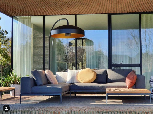 heizstrahler_von_heatsail_modell_disc_ueber_einer_gemuetlichen_outdoor-couch_mit_flachem_couchtisch_auf_der_ueberdachten_terrasse_eines_modernen_hauses_mit_grossen_glasfronten