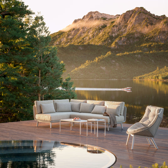 der_mbrace_sessel_von_dedon_neben_der_dedon_couchlandschaft_mbarq_und_zwei_couchtischen_auf_einer_hoelzernen_terrasse_mit_einem_pool_direkt_neben_einem_weiten_bergsee_im_hintergrund_mit_hohen_bewaldeten_bergen_dedon_gartenmoebel_im_onlineshop_henrik-bosse.de_erhaeltlich
