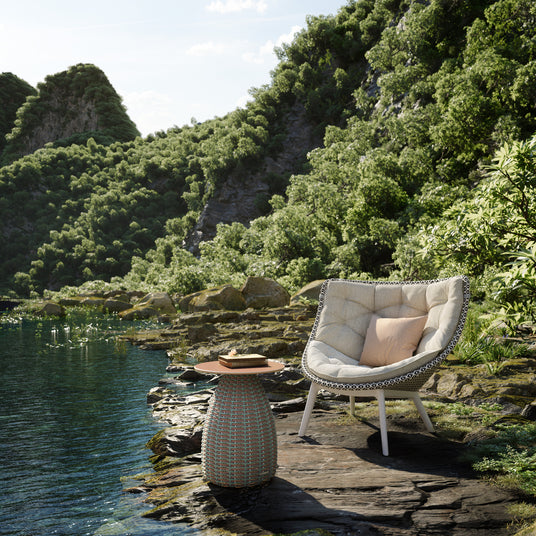 dedon_mbrace_sessel_und_dedon_porcini_beistelltisch_auf_einem_steinernen_ufer_an_einem_glitzernden_see_umgeben_von_gruen_bewachsenen_felsen_beides_im_onlineshop_henrik-bosse.de_erhaeltlich