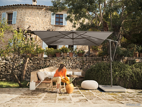 glatz_sunwing_casa_Ampelschirm_auf_einer_mediterranen_terrasse_vor_einem_grossen_haus_aus_stein_im_schutz_des_sonnenschirms_topft_eine_frau_eine_pflanze_ein