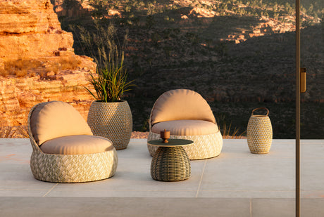 Das Bild zeigt zwei DEDON DALA Lounge-Sessel in hellem Geflecht, kombiniert mit einem passenden Beistelltisch und zwei grossen DALA Vasen. Die Szene ist auf einer Terrasse mit weitem Ausblick in eine rötlich leuchtende Berglandschaft aufgenommen. Weiches Abendlicht betont die sanften Rundungen der Möbel und ihre natürliche Einbindung in die Umgebung.